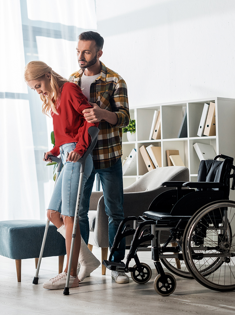 hoitaja auttaa loukkaantunutta naista, jolla on kainalosauvat pyörätuoliin